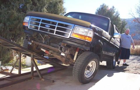 1996 Ford Bronco - A black 1990s Ford Bronco full-size SUV is driven front-first up a set of homemade steel ramps, with its front wheels elevated and undercar…