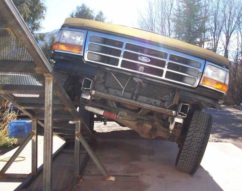 1996 Ford Bronco - Front 3/4 view of a green/tan 1990s Ford Bronco or F-series truck raised on metal ramps/stands, exposing the underside with visible front a…