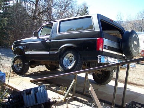 1996 Ford Bronco - A black two-door Ford Bronco SUV parked on an elevated metal ramp/lift, viewed from the rear passenger side, showing chrome rear bumper, a…