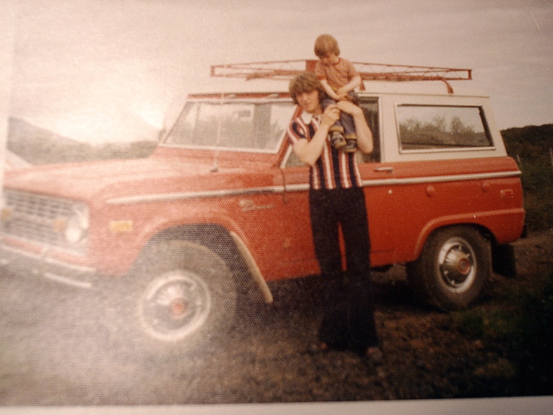 Vintage red and white Ford Bronco with roof rack; woman holds child beside it outdoors.
