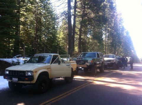 1989 Ford Bronco - A line of off-road vehicles, including a cream-colored Toyota pickup, are parked on a road in a forest setting.