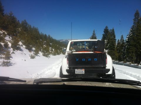 1989 Ford Bronco - A white Ford pickup truck drives through a snowy mountain pass, its tailgate clearly marked with the Ford logo.