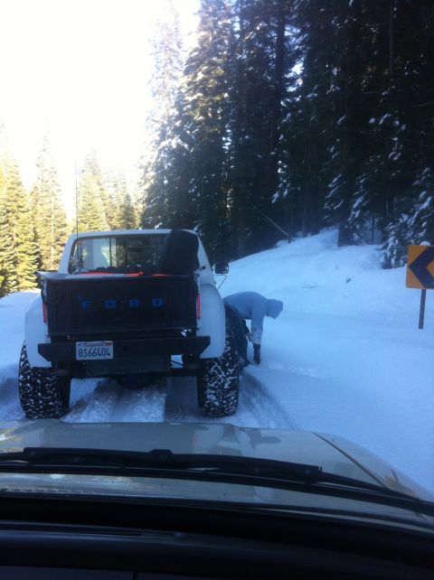 1989 Ford Bronco - A white Ford pickup truck with large tires is stuck in the snow, while a person attempts to clear it.
