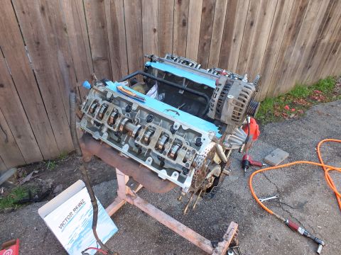 2003 Ford Crown Victoria - Engine block mounted on a repair stand against a wood fence, cylinder heads open and Victor Reinz gasket box on the ground.