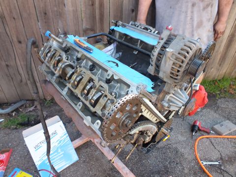 2003 Ford Crown Victoria - Disassembled V6 on a repair stand with alternator and chains exposed, Victor Reinz gasket box on the ground.