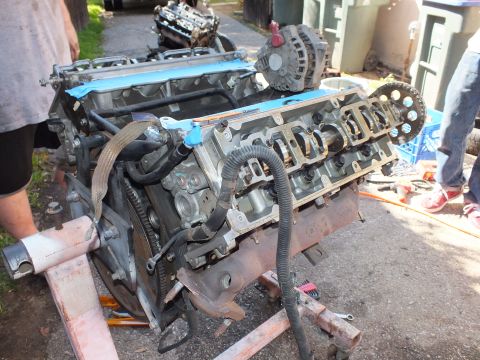 2003 Ford Crown Victoria - Foreground V6 with open heads and alternator beside a second engine, people standing around in a driveway.
