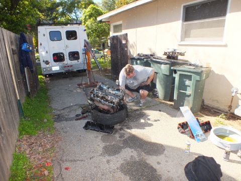 2003 Ford Crown Victoria - Wide shot of someone working on an engine balanced on a tire, white box truck and trash bins behind.
