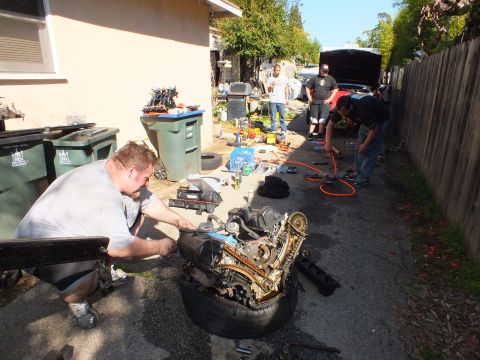 2003 Ford Crown Victoria - Wide driveway scene of several workers around engines, tools and a pickup with raised hood in the distance.