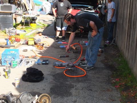 2003 Ford Crown Victoria - Person operating an air-powered tool over a part on the ground, orange hose coiled and others standing nearby.