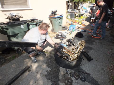 2003 Ford Crown Victoria - Worker tending an engine set on a tire near green bins, scattered tools and people down the driveway.