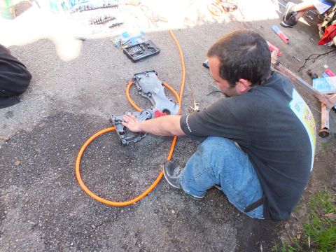 2003 Ford Crown Victoria - Person sitting on the ground working on a metal cover with tools and an orange hose around them.