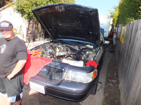 2003 Ford Crown Victoria - Gray Ford sedan with raised hood during engine work, red rags draped over the front and a CA plate.