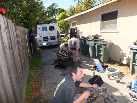 2003 Ford Crown Victoria - Driveway scene with a man in foreground holding a part and others over an engine by a van.