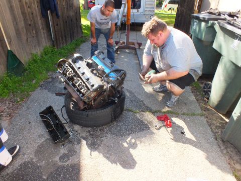 2003 Ford Crown Victoria - Removed engine balanced on a tire while two men inspect parts beside trash bins and a fence.