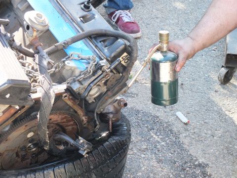 2003 Ford Crown Victoria - Hand-held brass torch cylinder pointed at a greasy engine block resting on a tire.
