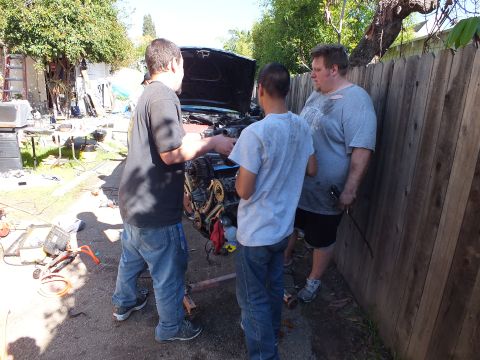 2003 Ford Crown Victoria - Men conversing beside a removed engine on a stand near a sedan with its hood open.