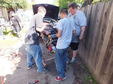 2003 Ford Crown Victoria - Group maneuvering a removed engine near the front of a sedan with the hood raised.