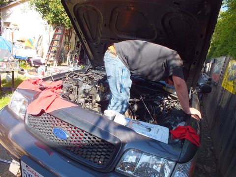 2003 Ford Crown Victoria - Mechanic reaching into the engine bay of a gray Ford SUV with red grille and raised hood.
