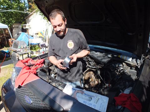 2003 Ford Crown Victoria - Mechanic inspecting a small part next to the exposed engine bay of a gray Ford.