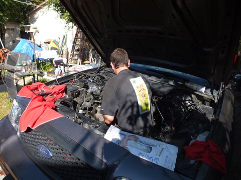 2003 Ford Crown Victoria - Mechanic working deep in the engine bay of a gray Ford with documents laid on the fender.
