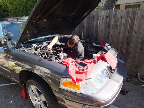 2003 Ford Crown Victoria - Mechanic working under the open hood of a gray Ford sedan with alloy wheels in evening light.