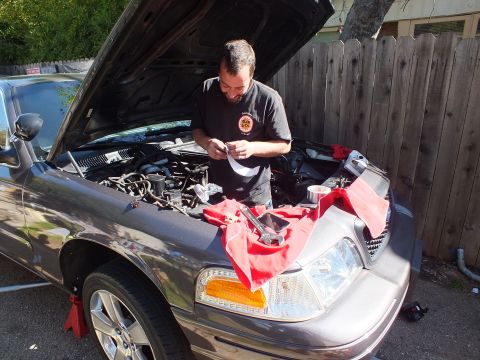 2003 Ford Crown Victoria - Mechanic reads paperwork over the open engine bay of a gray sedan, red shop towel draped on the fender.