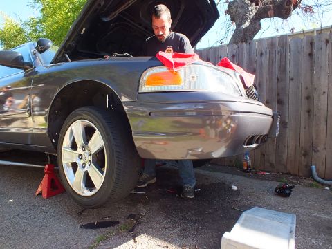 2003 Ford Crown Victoria - Gray sedan jacked up at the front while a person works near the bumper, scattered tools on pavement.
