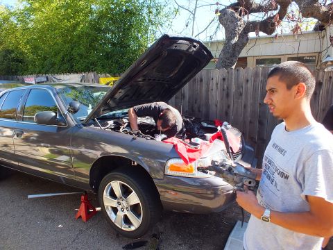2003 Ford Crown Victoria - One man leans into the engine bay of a gray sedan while another holds a removed part nearby.