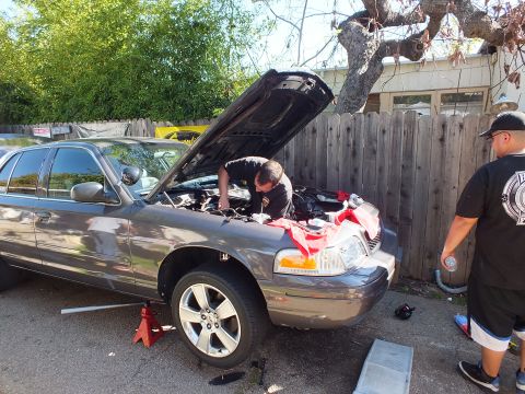 2003 Ford Crown Victoria - gray Crown Victoria sedan sits on a jack stand as two men work at the open hood by a fence.