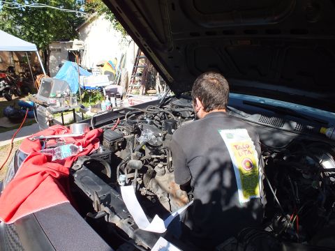 2003 Ford Crown Victoria - Worker reaches into a sedan engine bay; a red rag and Tang packet rest on the fender.