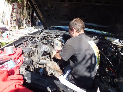 2003 Ford Crown Victoria - Worker reaches into the engine bay of a dark sedan in a tool-strewn yard, red rag on the fender.