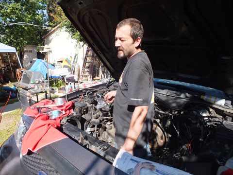 2003 Ford Crown Victoria - Worker leans over the engine bay of a dark sedan beside a wood fence, tent and clutter behind.