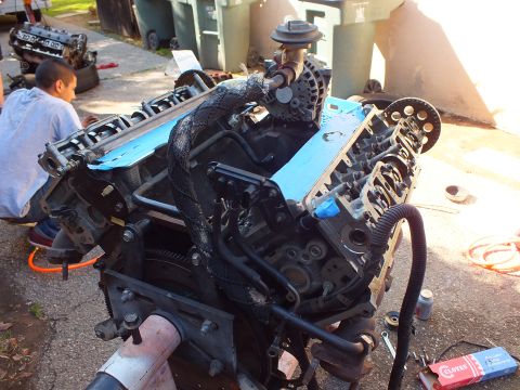 2003 Ford Crown Victoria - Disassembled V8 engine on a stand showing cam towers and timing gear, person crouched behind.