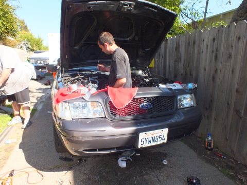2003 Ford Crown Victoria - Gray Ford Crown Victoria, hood up and plate 5YWN854, as a man works at the engine by a fence.