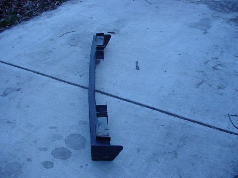 1992 Ford Bronco - A dark bumper is shown lying on a concrete surface with some leaves in the background.