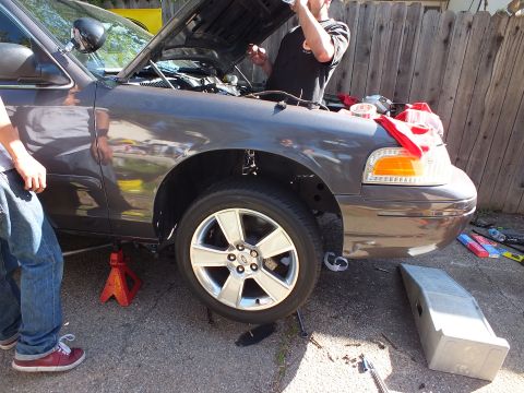 2003 Ford Crown Victoria - Gray Ford sedan jacked at the front, alloy wheel and amber marker visible, men working at the hood.