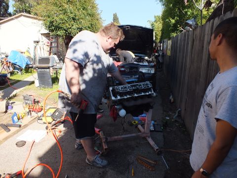 2003 Ford Crown Victoria - Worker uses a power tool on a V8 block mounted on a stand; a gray sedan sits behind in the alley.