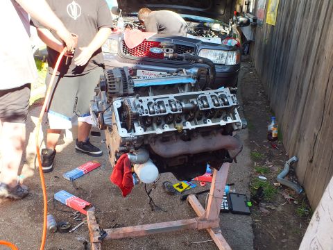 2003 Ford Crown Victoria - V8 engine on a stand with one head exposed and cam visible, gray Ford sedan with hood up behind.
