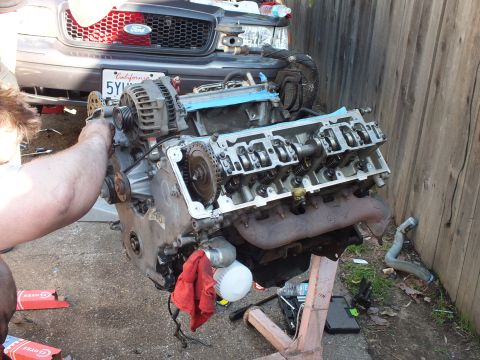 2003 Ford Crown Victoria - Hands work an exposed V8 valvetrain on a stand; gray Ford grille and CA plate 5YWN854 behind.