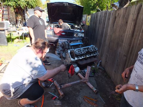 2003 Ford Crown Victoria - man kneels by a V8 engine on a stand in a narrow alley, gray Ford sedan with hood up behind.