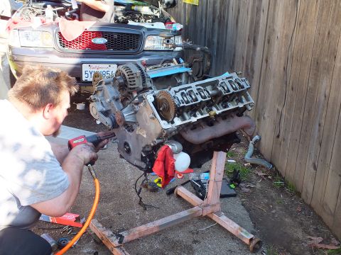 2003 Ford Crown Victoria - Worker drills into a V8 engine on a stand; gray Ford grille and CA plate 5YWN854 in background.