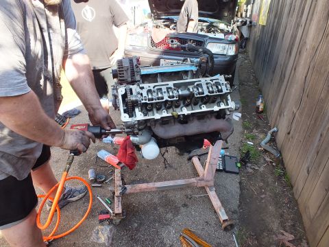 2003 Ford Crown Victoria - Worker runs an impact driver on a V8 on a stand in a narrow alley, tools scattered on the ground.