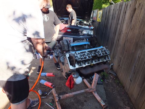 2003 Ford Crown Victoria - Two men work a V8 engine on a stand in a narrow alley as a gray Ford sedan sits behind, hood up.