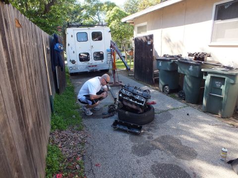 2003 Ford Crown Victoria - Man crouches by an engine resting on a tire in a driveway, white box van and green bins behind.