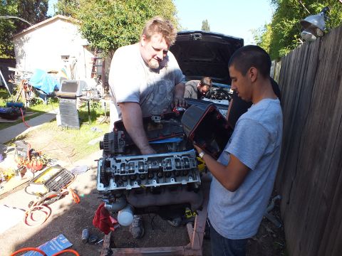 2003 Ford Crown Victoria - Backyard engine teardown: a man and a younger helper handle an exposed V8 cylinder head next to an open-hood Ford