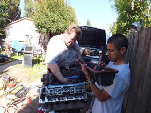 2003 Ford Crown Victoria - Two people inspect a red intake component while a V8 engine sits on a stand along a wood fence