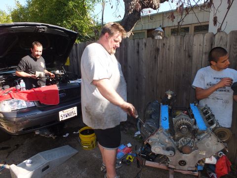 2003 Ford Crown Victoria - Group rebuild scene: one man pours fluid into a gray Ford while another works a V8 on a stand, CA plate 5YWN854