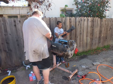 2003 Ford Crown Victoria - V8 engine rests on a homemade timber stand as two workers handle a part along the fence