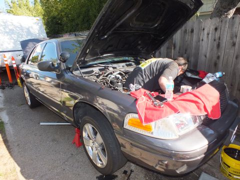 2003 Ford Crown Victoria - person leans into the engine bay of a gray Ford sedan, hood up and fender draped with a red towel