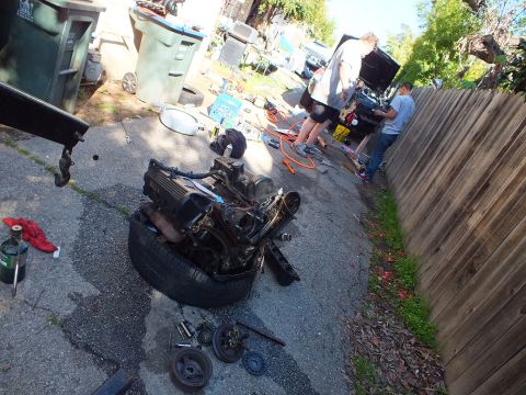 2003 Ford Crown Victoria - pulled V8 engine sits on a tire amid scattered parts while work continues on a car down the driveway